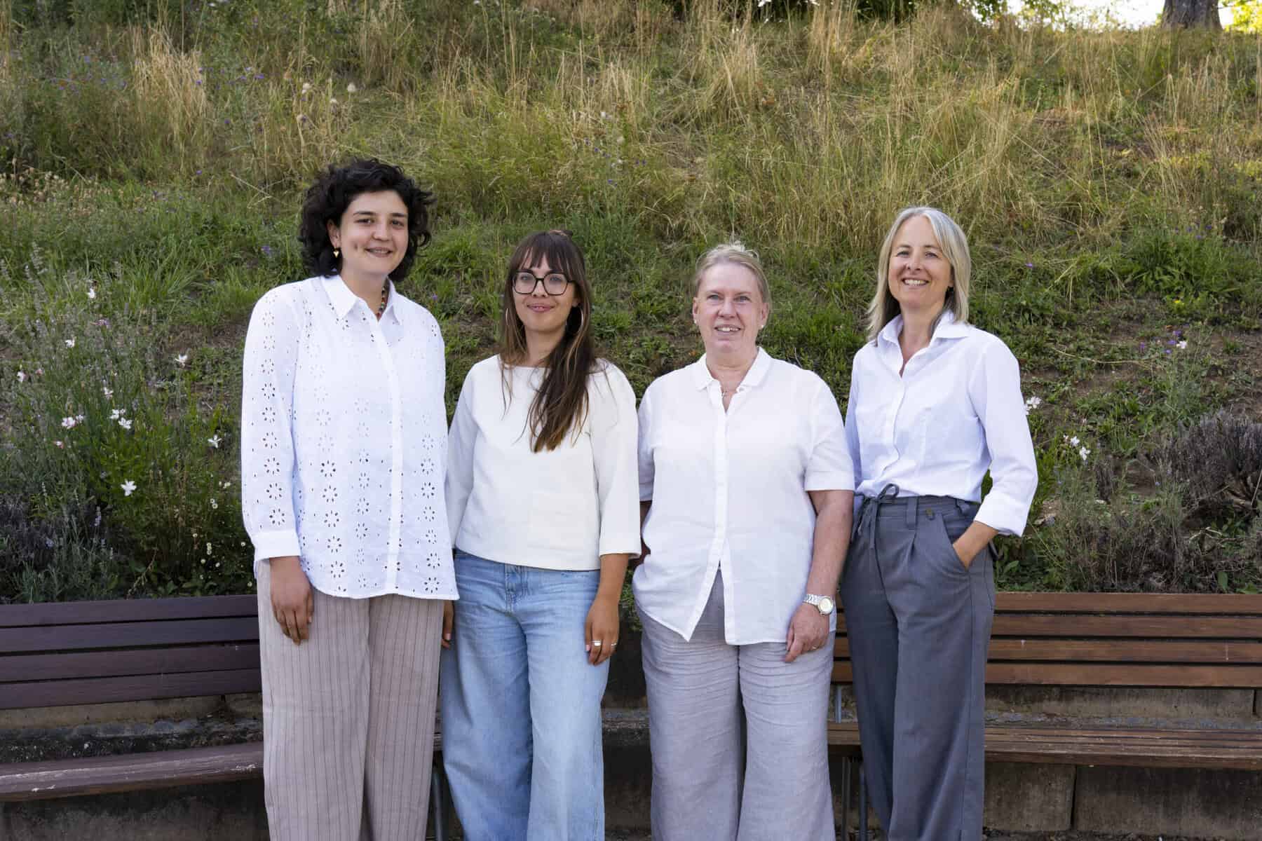 Auf dem Bild sind 4 Personen zu sehen, sie stehen vor Bänken im freien. Sie tragen alle weiße Hemden und lächeln.