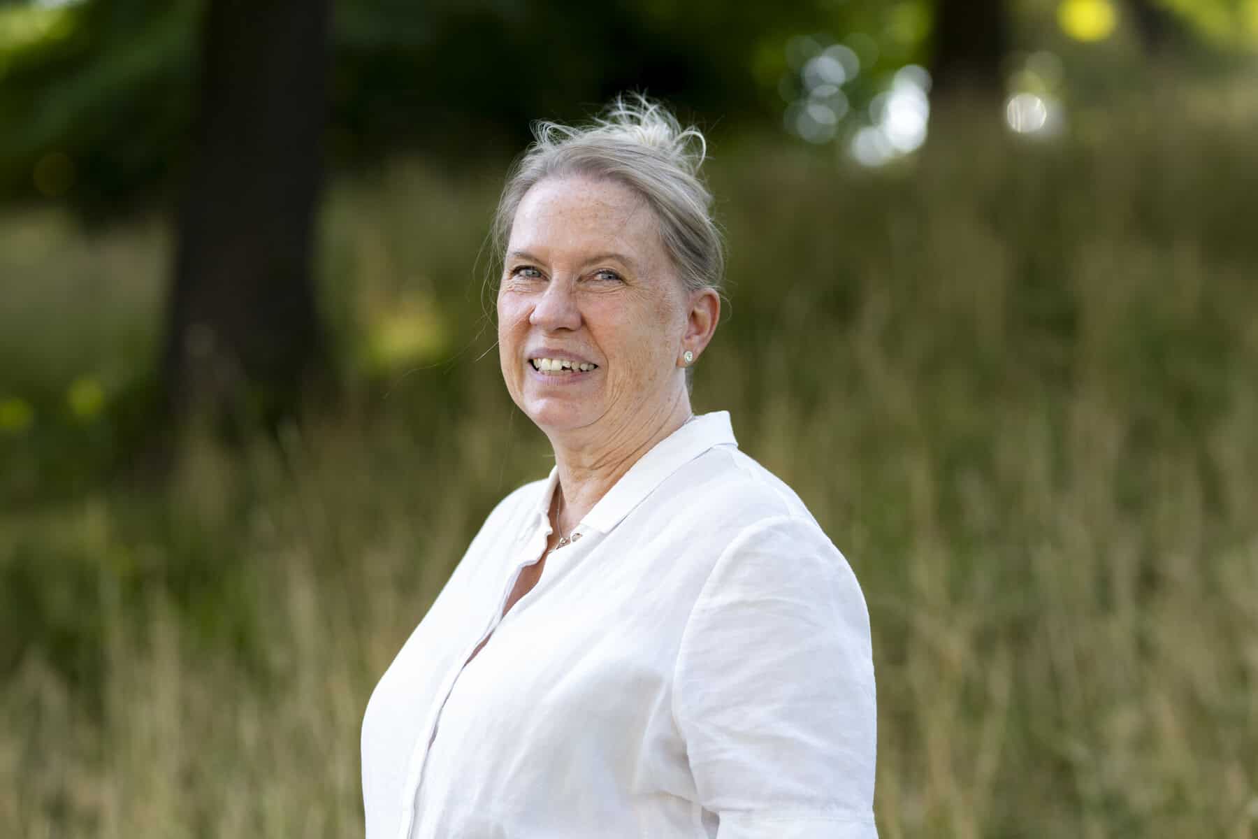 Bild von einer Person mit zurückgebundenen hellen Haaren und weißem Oberteil vor einer grünen Wiese und Bäumen.