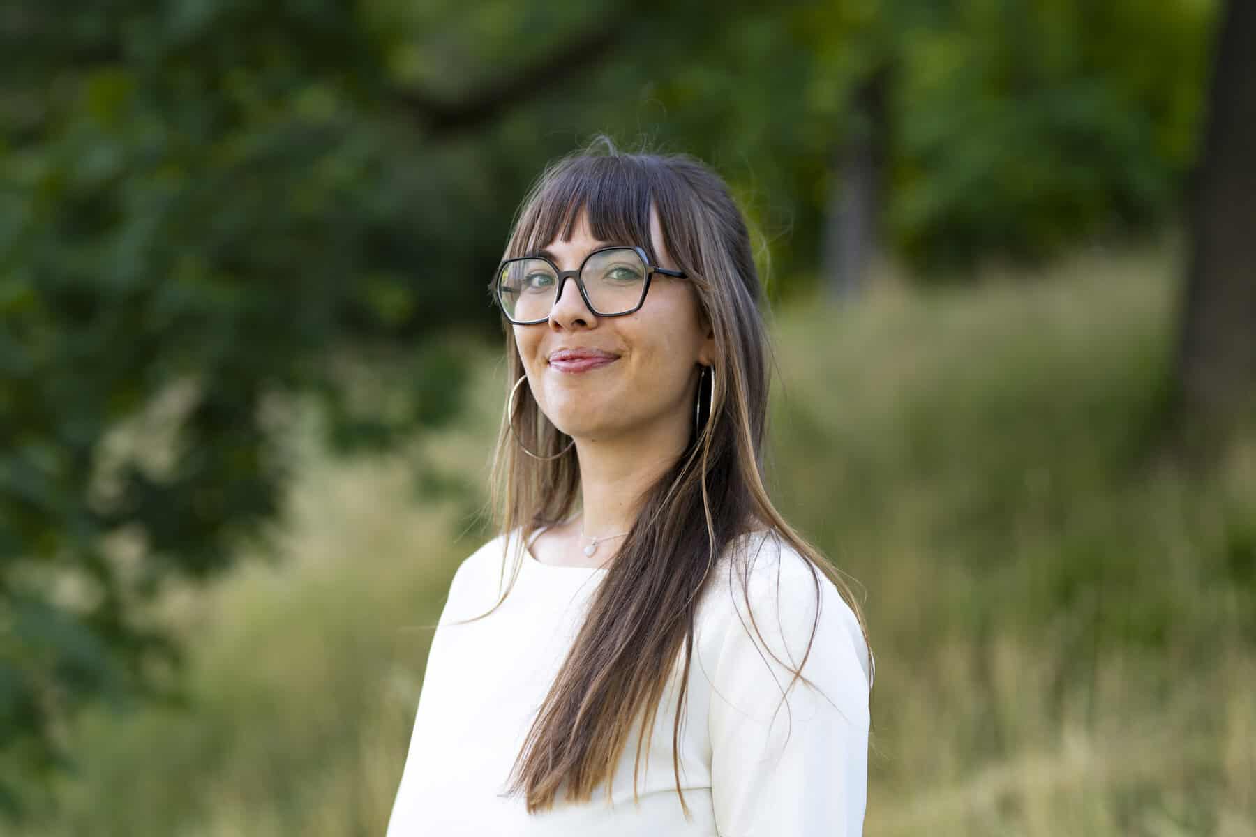 Eine Person steht im Freien vor Bäumen und Wiese. Die Person trägt ein helles Oberteil und eine Brille. Im Hintergrund sind grüne Pflanzen zu sehen