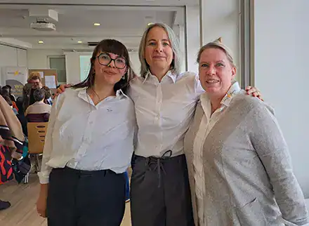 Drei Frauen nebeneinander. Sie tragen weiße Hemden. Die mittlere Frau hat ihre Hände auf den Schultern der beiden Frauen neben ihr.