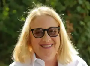 Porträtfoto einer Frau mit kurzen blonden Haaren. Sie trägt ein weißes Hemd. Sie trägt eine Brille. Sie lächelt in die Kamera.