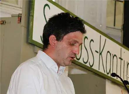 Ein Mann in einem weißen Hemd. Er steht an einem Rednerpult. Er schaut nach unten.