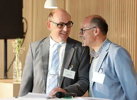 Zwei Männer in Anzügen an einem Stehtisch. Sie haben beide ein Namensschild an ihrem Anzug. Sie reden miteinander.