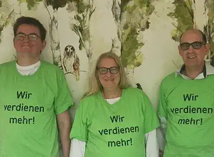 Drei Menschen stehen nebeneinander. Zwei Männer und in der Mitte eine Frau. Sie tragen alle ein grünes T-Shirt mit der Aufschrift "Wir verdienen mehr!".