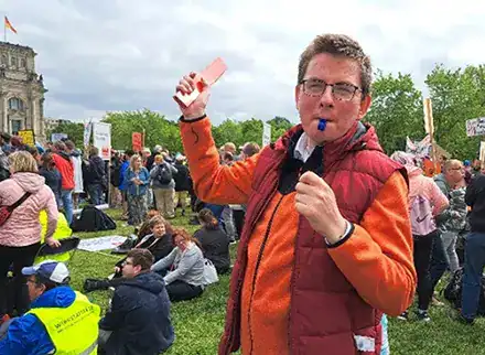 Ein Mann hält ein Stück Karton in seiner Hand. Im Mund hat er eine Trillerpfeife. Im Hintergrund sind viele Menschen die demonstrieren.
