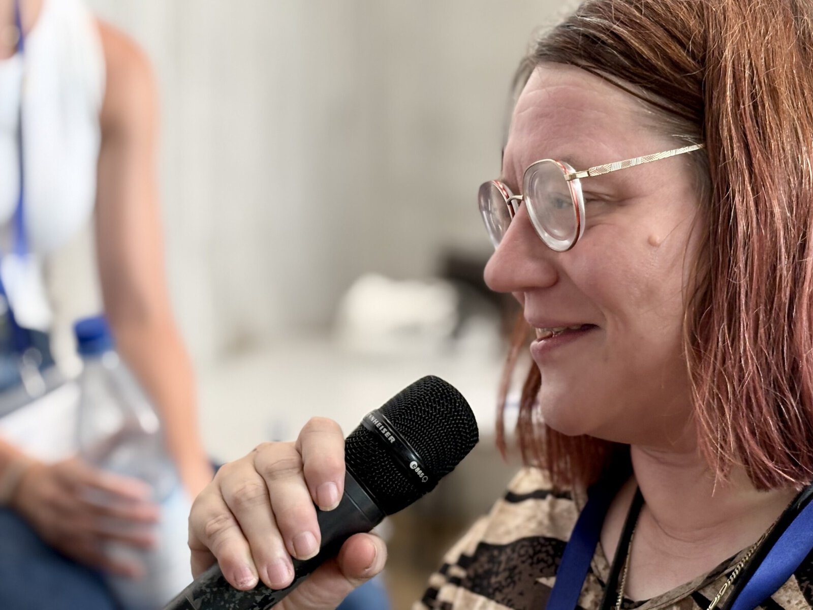 Auf dem Bild ist eine Person zu sehen, die in ein Micro spricht. Das Bild ist sehr nah aufgenommen. Sie hat rote Haare und eine Brille.
